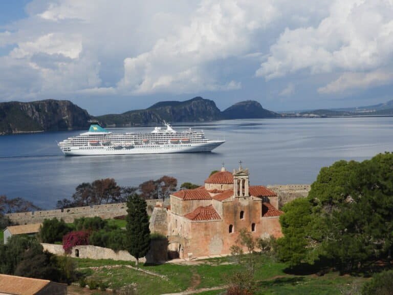 Cruise ship sailing near historic coastal village with church and lush greenery.