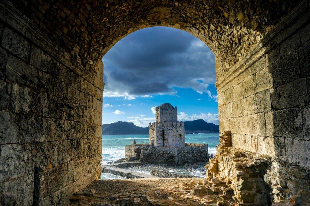 Coastal fortress viewed through stone archway with sea and mountains in background.
