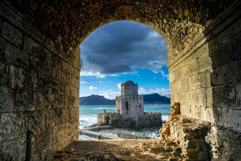 Coastal fortress viewed through stone archway with sea and mountains in background.