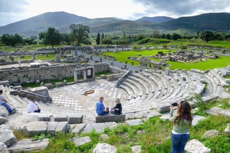 Open-air ancient Greek amphitheater with visitors and scenic mountain backdrop.