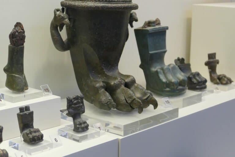 Ancient artifacts including a large bronze vessel with a foot-shaped base, displayed in a museum set.