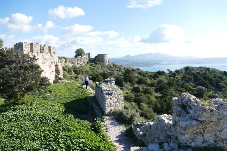 Ancient ruins overlooking a lush green landscape with mountains and water in the background.