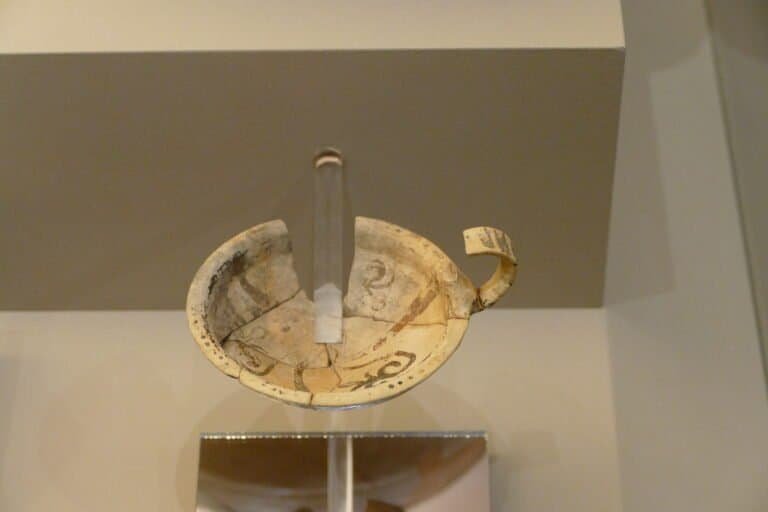 Antique Greek oil lamp with a wick and handle, displayed on a white surface.