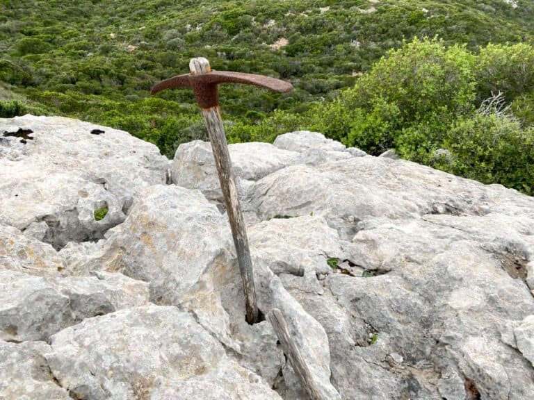 Old pickaxe embedded in rocky terrain with green shrubbery in the background.